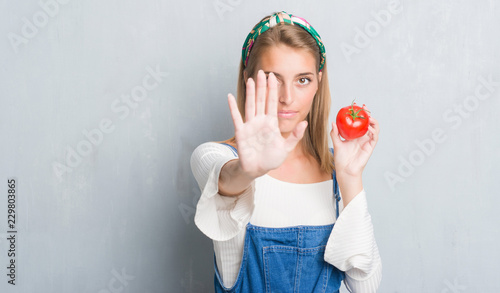 Beautiful young woman over grunge grey wall eating fresh tomato with open hand doing stop sign with serious and confident expression  defense gesture