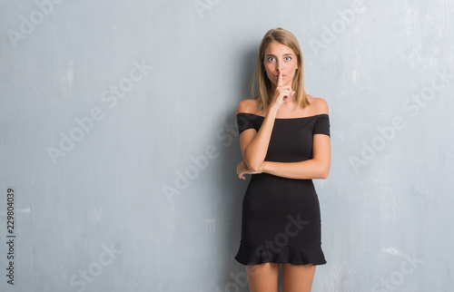 Beautiful young woman standing over grunge grey wall wearing elegant dress asking to be quiet with finger on lips. Silence and secret concept. © Krakenimages.com