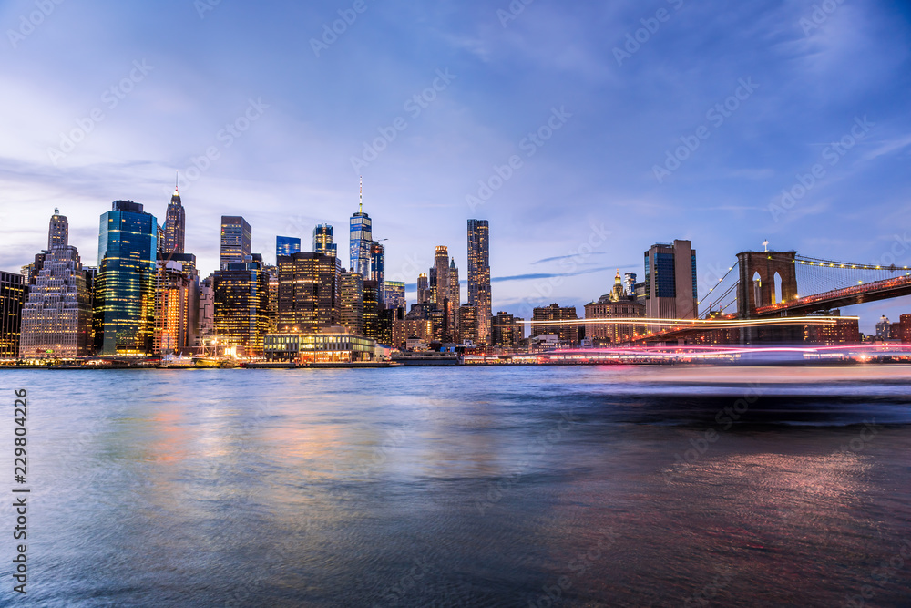 Obraz premium Widok z zewnątrz na Nowy Jork Brooklyn Bridge Park w Nowym Jorku nad wschodnią rzeką, panoramę miasta o zachodzie słońca, zmierzch, zmierzch, niebieska godzina, ciemna noc, drapacze chmur, budynki, fale, smugi świetlne łodzi wycieczkowej
