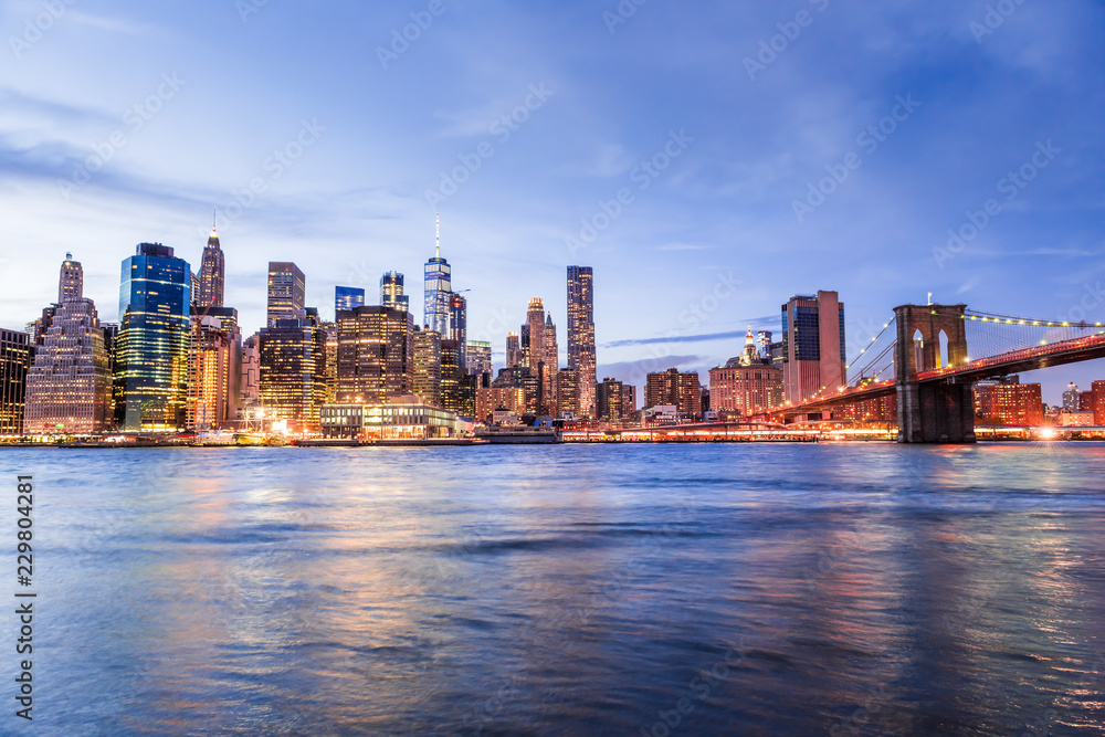 Wide angle view on NYC New York City Brooklyn Bridge Park by east river, cityscape skyline at dusk, twilight, blue hour, dark night, skyscrapers, illuminated, wave, reflection, bright light