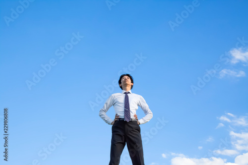 青空をバックに腰に両手を当て見上げる若いYシャツ姿の男性1人立ち姿。ビジネス、成功、野心、向上心イメージ © chikala