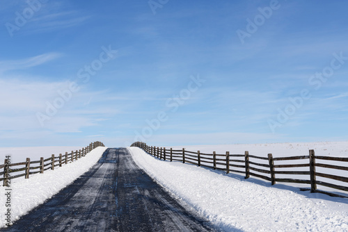 Fototapeta Naklejka Na Ścianę i Meble -  Empty Road and Snow Covered Fields - looking up small hill and bordered by wooden fence