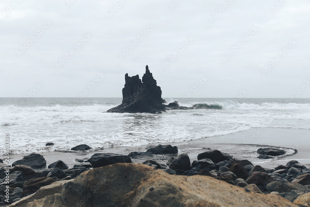 lonely sharp rock in the Atlantic ocean with stones on the beach and black sand in the cloudy evening Benijo, Tenerife, Spain