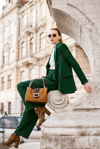 Outdoor full body fashion portrait of fashionable woman wearing sunglasses, white turtleneck, green suit, blazer, trousers, leopard print boots, holding suede bag, posing in street of european city