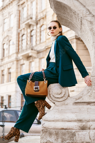 Outdoor full body fashion portrait of young woman wearing sunglasses, white turtleneck, blue suit, blazer, trousers, leopard print ankle boots, holding suede bag, posing in street of european city