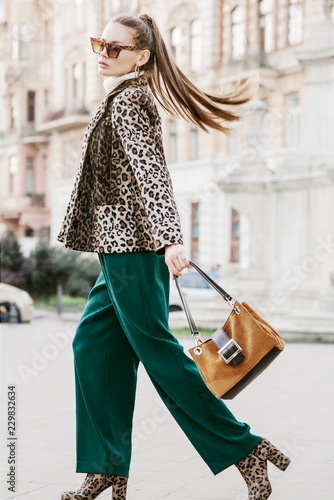 Outdoor full body fashion portrait of young beautiful fashionable woman wearing sunglasses, leopard print blazer, boots, green trousers, holding brown suede bag, walking in street of european city
