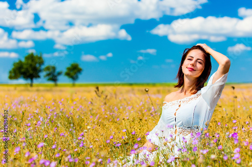 photo of the beautiful young woman in white dress sitting in the field in summer time