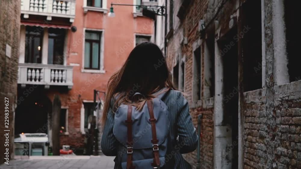 Camera follows young female tourist with backpack walking along beautiful dark ancient city street in Venice slow motion Camera follows young female tourist with backpack walking along beautiful dark ancient city street in Venice slow motion