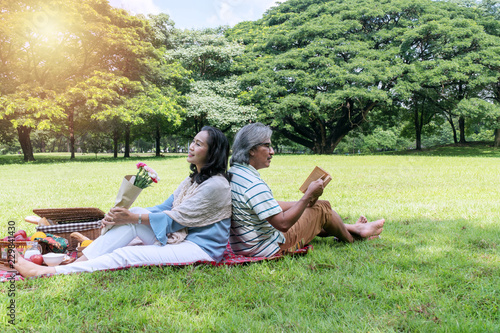 Elderly lifestyle concept. Happiness senior couple sitting back to back relax together in the public park. Husband reading book his wife holding flower with picnic on green grass.