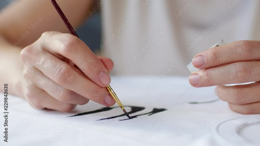Close up shot of hands of unrecognizable female artist creating hand painted design on white t-shirt using acrylic paint Close up shot of hands of unrecognizable female artist creating hand painted design on white t-shirt using acrylic paint