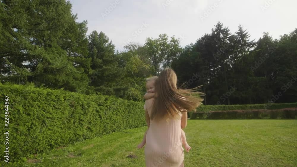 Slow motion of baby girl with mother running away on green grass in the summer light in park. Baby have fun. Baby laughing. Green thuja background. Happy family concept. Woman with long hair. Slow motion of baby girl with mother running away on green grass in the summer light in park. Baby have fun. Baby laughing. Green thuja background. Happy family concept. Woman with long hair.