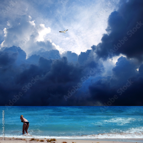 Young adult woman in bikini relaxing on sandy beach over cloudy storm sky with flying seagull