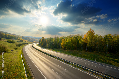 Fototapeta Empty asphalt highway between deciduous forest in autumn colors under the radiant sun and dramatic clouds