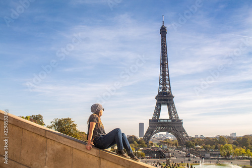 Canvas Print Young ginger woman looking at the Eiffel Tower sitting by the Trocadero stairs on a beautiful day of summer with blue sky and clouds in Paris, France