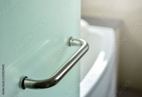 Glass door handle of a glass partition shower unit. Bathroom glass door detail with bath tub in the background. Selective focus and soft light effect added.