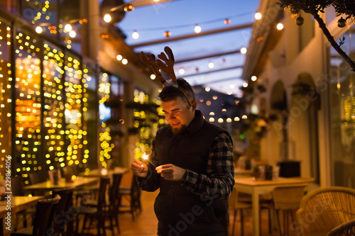 Christmas, people and winter holidays concept - surprised man in horns of deer standing at night street with bengal lights in his hands