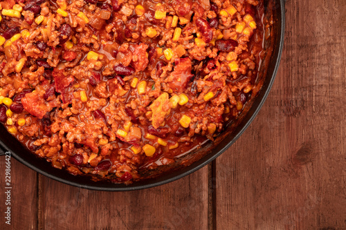A closeup photo of a large cast iron pan with chili con carne, shot from the top on a dark rustic wooden background with a place for text