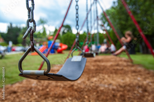 Fototapeta Naklejka Na Ścianę i Meble -  Closeup picture of a swing in a park for kids. Kids swigning in the blurry background - Picture taken on a warm summer day, with mulch ground instead of sand, and green lawn in the background