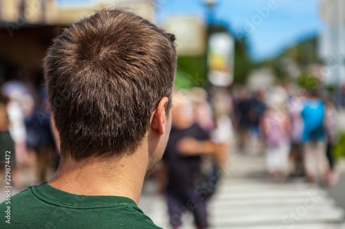 Young man with a green shirt is looking forward before crossing the street, with many blurred people in the background - Shot from the back during an alternative political gathering