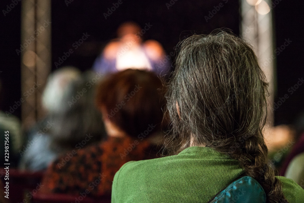 Man with long hairs and wearing a green shirt takes part of an audience and listens to a public conference given in a church - Pictured from the back with blurry people in the background