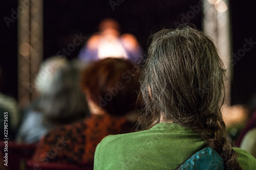 Man with long hairs and wearing a green shirt takes part of an audience and listens to a public conference given in a church - Pictured from the back with blurry people in the background photo