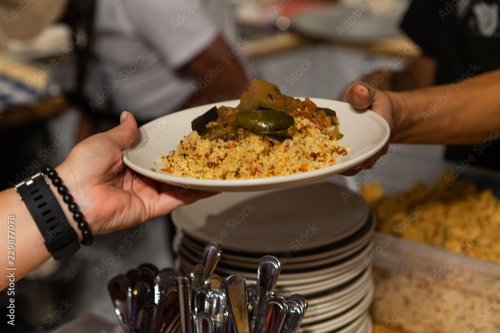 Woman wearing a watch and a black beads bracelet is picking up a plate full of couscous and ratatouille - With a pile of plates and utensils in the background