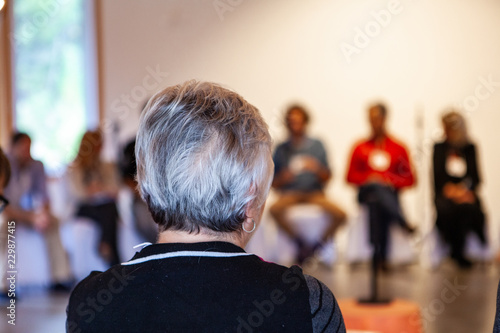 Woman with short black and white hairs is talking in front of a full committee - Pictured from the back in an alternative health center photo