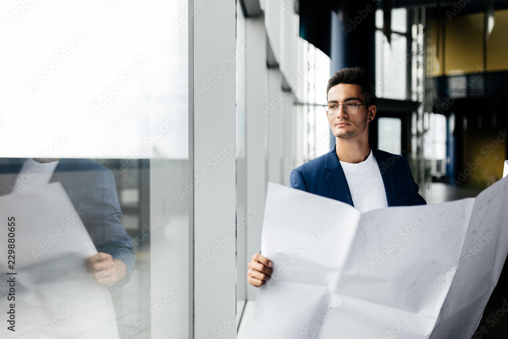 Architect in stylish clothes holds sheet with drawing in his hand and talks by phone on the background of a modern glass multistory building Architect in stylish clothes holds sheet with drawing in his hand and talks by phone on the background of a modern glass multistory building