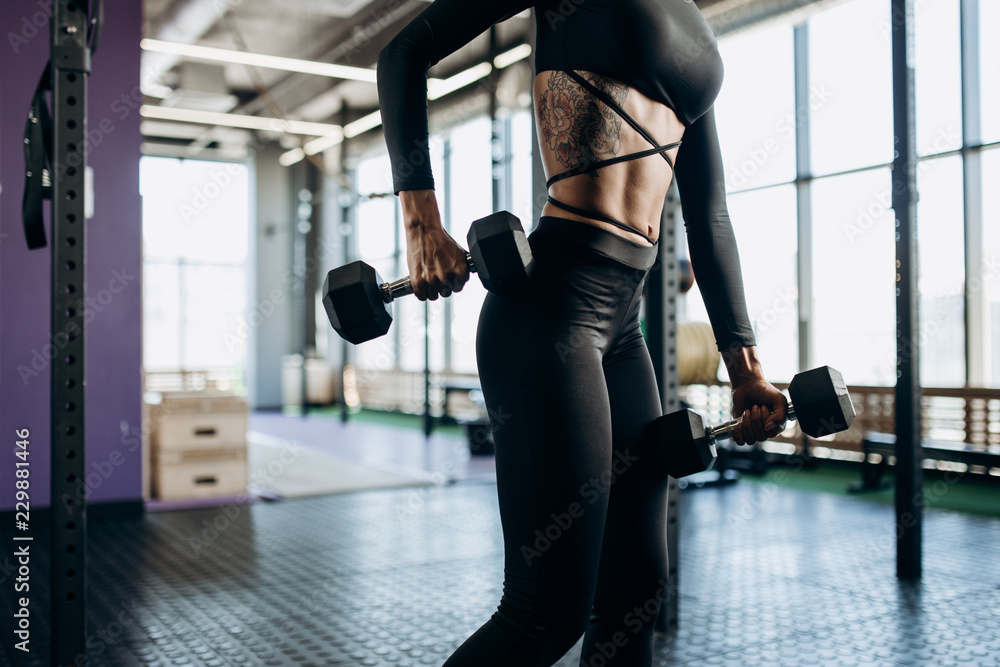 Slender body of a young woman with tattoo in a black sportswear that is doing exercises with dumbbells in the gym Slender body of a young woman with tattoo in a black sportswear that is doing exercises with dumbbells in the gym