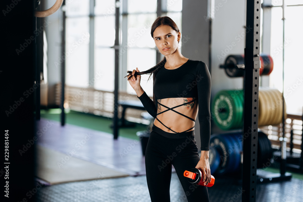 Athletic dark-haired girl dressed in black sportswear stands with water in her hand near the sport equipment in the gym and touches hair
