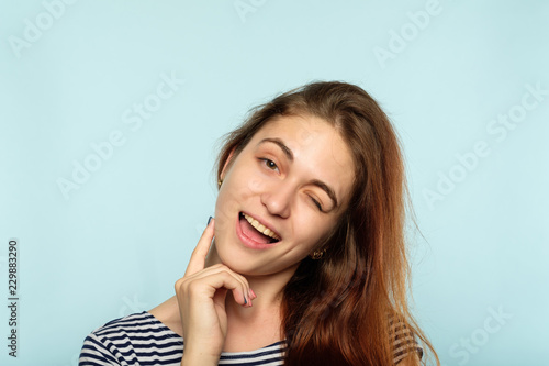 emotional facial expression. smiling woman pleased with herself. young beautiful brown haired girl winking. portrait on blue background.