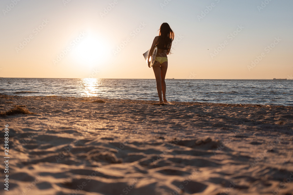 Dark-haired girl in a swimsuit stands on the sandy beach near the sea on the sunset and holds a surfboard