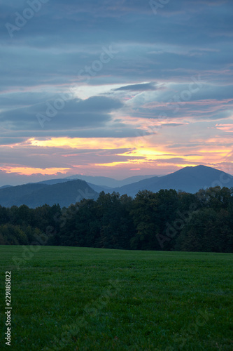 Cloudy sunrise on the green meadow, dark forest and Beskydy mountains in the distance