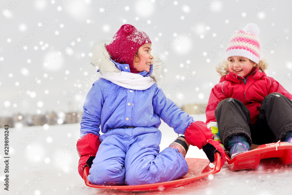 childhood, sledging and season concept - happy little girls on sleds outdoors in winter