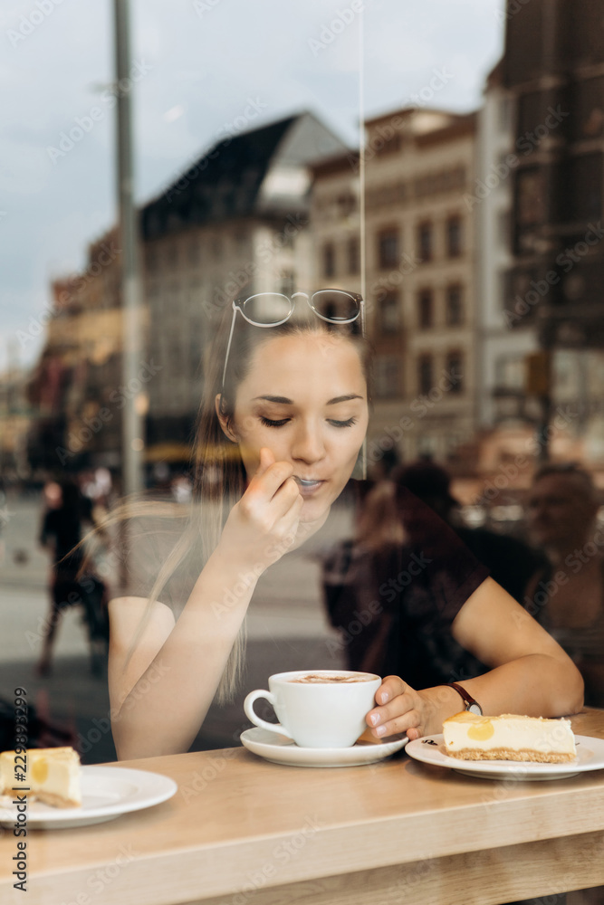 View through the window. A young girl eats a cheesecake or dessert and drinks coffee in a cafe in Leipzig in Germany.