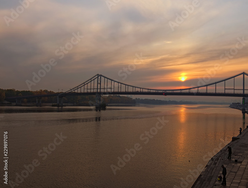 Pedestrian Bridge over Dnipro River during sunrise. The sun is reflected in the water. Men are fishing on the embankment. Autumn landscape. Kyiv, Ukraine