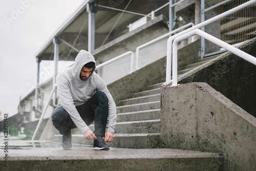 Sporty man getting ready for winter running and outdoor fitness workout under the rain. Sportsman lacing sport shoes on urban stairs.