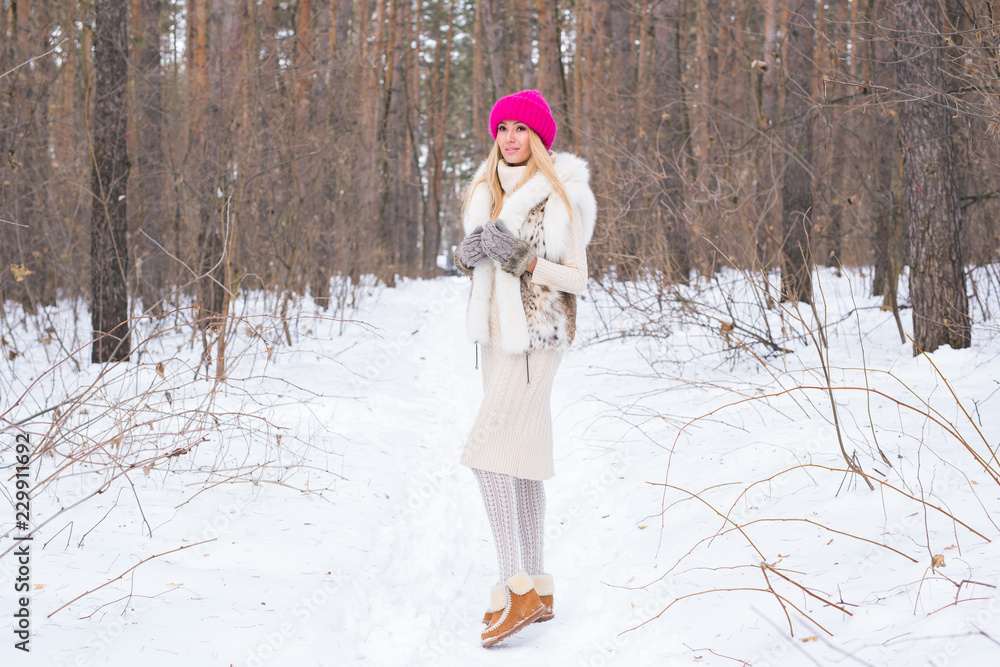 Season and people concept - Attractive blond woman dressed in white coat and pink hat standing in winter snowy park Season and people concept - Attractive blond woman dressed in white coat and pink hat standing in winter snowy park