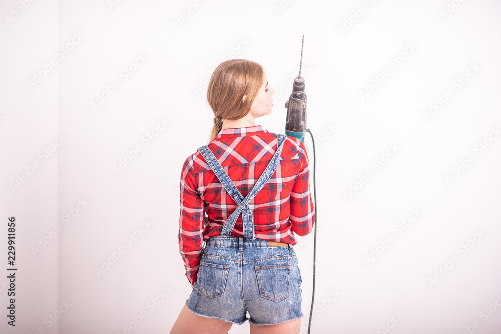 A young sexy woman stands with her back near the white wall and holds a drill. The girl in shorts and a plaid shirt makes repairs using a perforator. woman makes repairs with a tool. A young sexy woman stands with her back near the white wall and holds a drill. The girl in shorts and a plaid shirt makes repairs using a perforator. woman makes repairs with a tool.