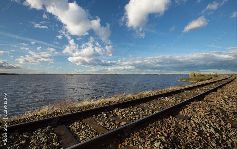 Railway tracks running along the lake with blue sky and white clouds