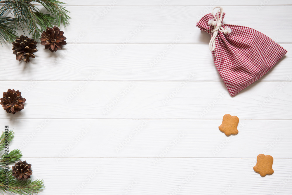 Obraz premium Christmas holidays background, copy space. Fir and pine tree branches with pinecones, christmas chequered red and white bags, ginger cookies on white wooden background, top view.