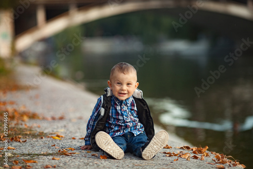Fototapeta Naklejka Na Ścianę i Meble -  A little boy is sitting next to the yellow autumn leaves and lake during a walk in the park.