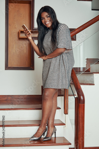 Side view of cheerful trendy black woman in dress and heels standing on wooden stairs of house with phone smiling at camera