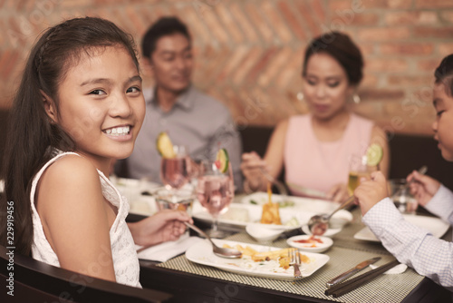 Portrait of pretty smiling girl sitting at the table with her family during dinner at restaurant