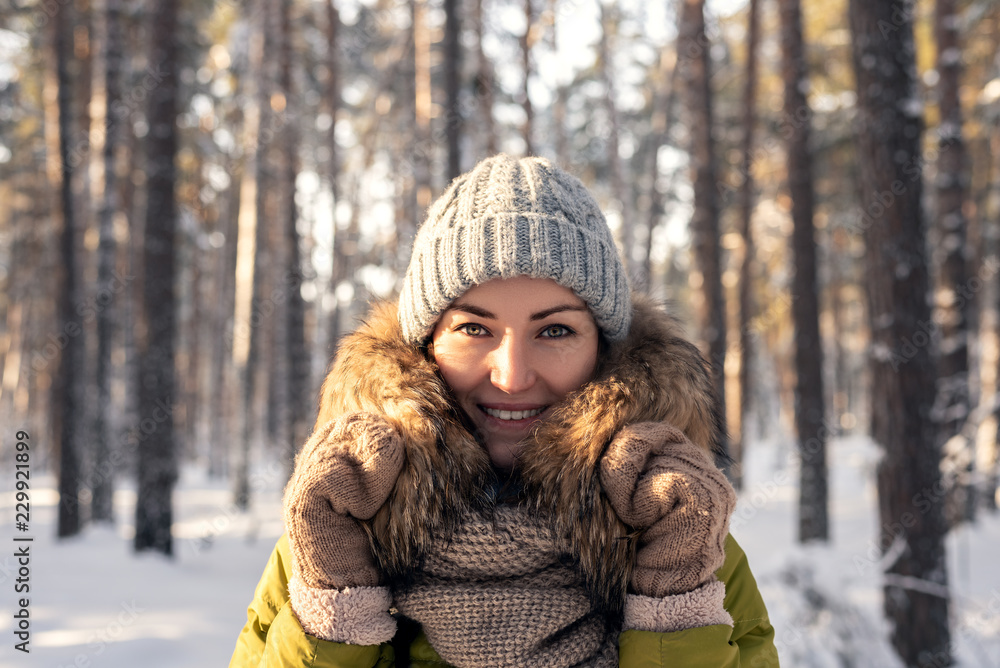 Horizontal winter portrait of a young woman on a coniferous forest background on a Sunny day. Girl in a jacket with fur, knitted hat and mittens smiling at the camera