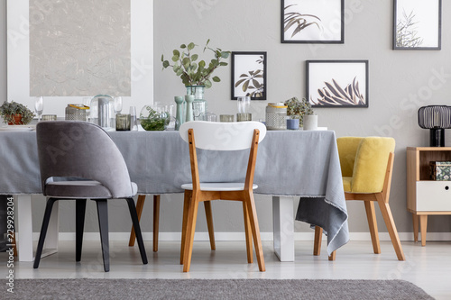 Modern chairs at table with tableware in grey dining room interior with posters and plants. Real photo