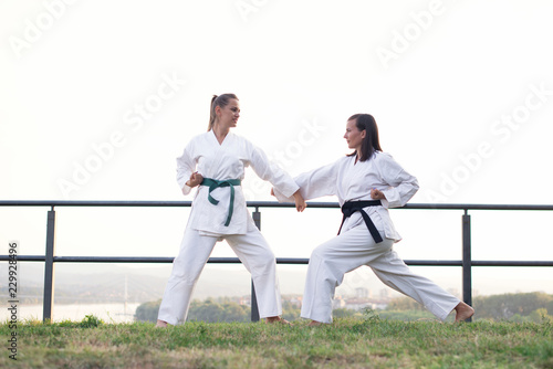 Two fit young women dressed in white kimono training karate martial arts in nature.