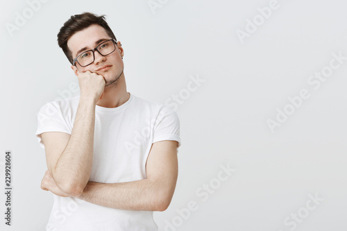What boring day at work. Portrait of indifferent apathic and unintersted attractive young man in glasses leaning head on hand from boredom looking at camera with no interest and joy over grey wall photo