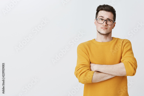 Displeased guy waiting for explanations. Confused doubtful and dissatisfied young handsome caucasian male in yellow sweater and glasses crossing hands on chest in hesitation and uncertainty photo