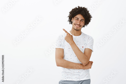 Door is there. Portrait of friendly happy male photographer in stylish t-shirt with afro hairstyle and moustache, tilting head and smiling broadly while pointing at upper left corner over grey wall